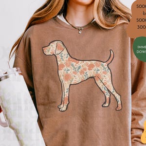Puede incluir: Una sudadera marrón con un diseño floral de perro. La ilustración del perro tiene un fondo crema y elementos florales naranjas, azules y verdes. La sudadera tiene cuello redondo y mangas largas.