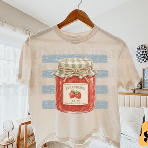 Op de afbeelding: Een crèmekleurig t-shirt met een afbeelding van een rode pot aardbeienjam met een groen-wit geblokt deksel vastgebonden met touw. De pot is gecentreerd over een achtergrond van horizontale lichtblauwe, verweerde strepen op de stof.
