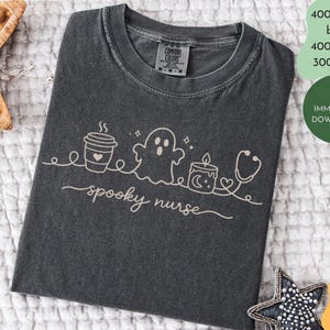 Könnte beinhalten: Ein anthrazitfarbenes T-Shirt mit Rundhalsausschnitt und einer hellen Strichgrafik, die eine Kaffeetasse, ein Gespenst, eine Kerze und ein Stethoskop zeigt. Der Text "spooky nurse" ist in Schreibschrift unter den Symbolen gedruckt. Comfort Colors Etikett.