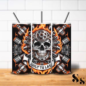 Puede incluir: Un juego de tres vasos de acero inoxidable con un diseño de calavera. La calavera está hecha de engranajes y está rodeada de llamas. Los vasos son negros con llamas naranjas y el texto "Built to Last".