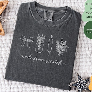 Könnte beinhalten: Dunkelgraues T-Shirt mit einer weißen Illustration einer Schleife, eines Glases mit Kräutern, einer Teigrolle und eines Blumenstraußes. Die Worte "made from scratch" sind kursiv geschrieben. Das Shirt ist aus Comfort Colors Material.