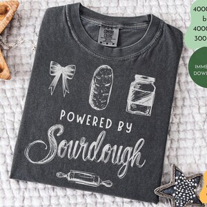 May include: Dark grey t-shirt featuring white illustrations of a bow, a loaf of bread, and a jar. The text "POWERED BY SOURDOUGH" is written in white script. A rolling pin illustration is at the bottom.
