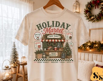 Puede incluir: Una camiseta de manga corta de color crema con un gráfico de un puesto de mercado navideño. El diseño incluye el texto "HOLIDAY MARKET," "MERRY MARKET," "FRESH GREENS," y "MERRY MARKET WREATHS COCOA GIFTS" junto con ilustraciones de coronas, cacao y regalos.