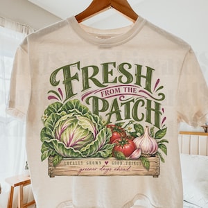 May include: A cream-colored short-sleeve t-shirt featuring a graphic of a wooden crate filled with a cabbage, tomatoes, and garlic. The text reads "FRESH FROM THE PATCH" in large green letters, with "LOCALLY GROWN" and "greener days ahead" below.