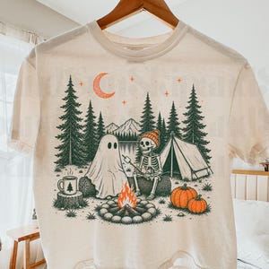 Może przedstawiać: Kremowy t-shirt z nadrukiem graficznym przedstawiającym ducha i szkielet siedzących na kłodzie przy ognisku. Scena zawiera namiot, sosny, góry, dwie dynie, kubek oraz pomarańczowy półksiężyc na nocnym niebie.