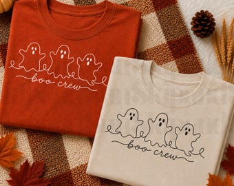 Puede incluir: Dos camisetas de cuello redondo, una en color naranja quemado y otra en crema, cada una con un gráfico de líneas blancas o negras de tres fantasmas conectados por una línea continua sobre el texto cursivo "boo crew" en la parte frontal.
