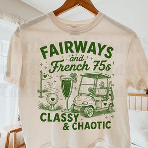 以下が含まれることがあります： クリーム色のTシャツにグリーンのグラフィックデザイン。デザインには、ゴルフカート、ゴルフボール、カクテルのイラストの上に「FAIRWAYS and French 75s」という言葉と、「CLASSY & CHAOTIC」という言葉が含まれています。
