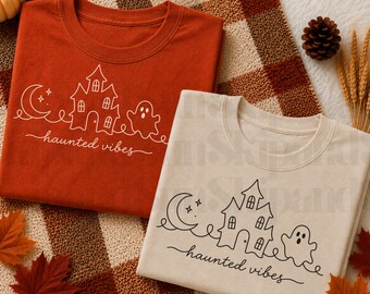 Puede incluir: Dos camisetas de manga corta, una en color naranja quemado y otra en crema. Ambas presentan un dibujo de línea continua de una luna creciente, una casa encantada y un fantasma, con el texto "haunted vibes" escrito en cursiva bajo el gráfico.