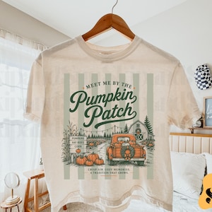 Op de afbeelding: Een crèmekleurig t-shirt met een grafische print met de tekst "Meet me by the Pumpkin Patch". Het ontwerp bevat een vintage oranje pick-up truck, een houten bord met "Pumpkins, Hay Rides, Cider" en een schuur, met de tekst "Crisp air, cozy memories, a tradition that grows."
