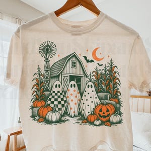 May include: A cream-coloured short-sleeve t-shirt featuring a graphic print of three ghosts standing in front of a barn, a windmill, and corn stalks. The design includes orange pumpkins, a crescent moon, and bats in a green and orange colour palette.