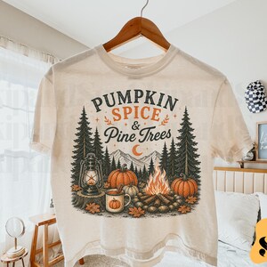 Puede incluir: Una camiseta de color crema con un estampado gráfico de una escena forestal con montañas, pinos, una fogata, calabazas, una linterna y una taza. El texto "PUMPKIN SPICE & Pine Trees" está impreso en negro y naranja sobre la ilustración.