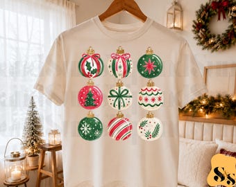 Puede incluir: Una camiseta de manga corta color crema con un estampado gráfico de nueve adornos navideños distintos dispuestos en cuadrícula. Los adornos muestran varios patrones festivos que incluyen acebo, un pino, copos de nieve, rayas y estrellas en rojo, verde y blanco.