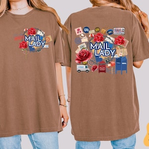 May include: Two brown t-shirts with a mail theme. The front of one shirt has a graphic of a globe with the text "Mail Lady". The back of the other shirt has a graphic of a mail truck, mailboxes, and roses with the text "Mail Lady".