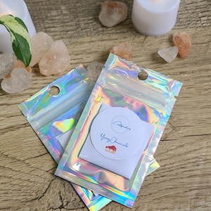 Może przedstawiać: Dwie iridiscentne, holograficzne torebki strunowe z białą okrągłą naklejką w środku, z tekstem "Citygardens" i "Young Cheesecake". Torebki leżą na drewnianej powierzchni, z białą świecą, kryształami i małymi kamieniami w tle.