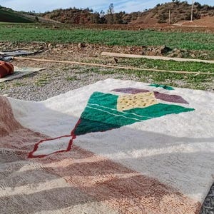 May include: A white, handwoven rug with a geometric design in shades of green, brown, and red. The rug is laid out on a gravel surface with a green field and blue sky in the background.