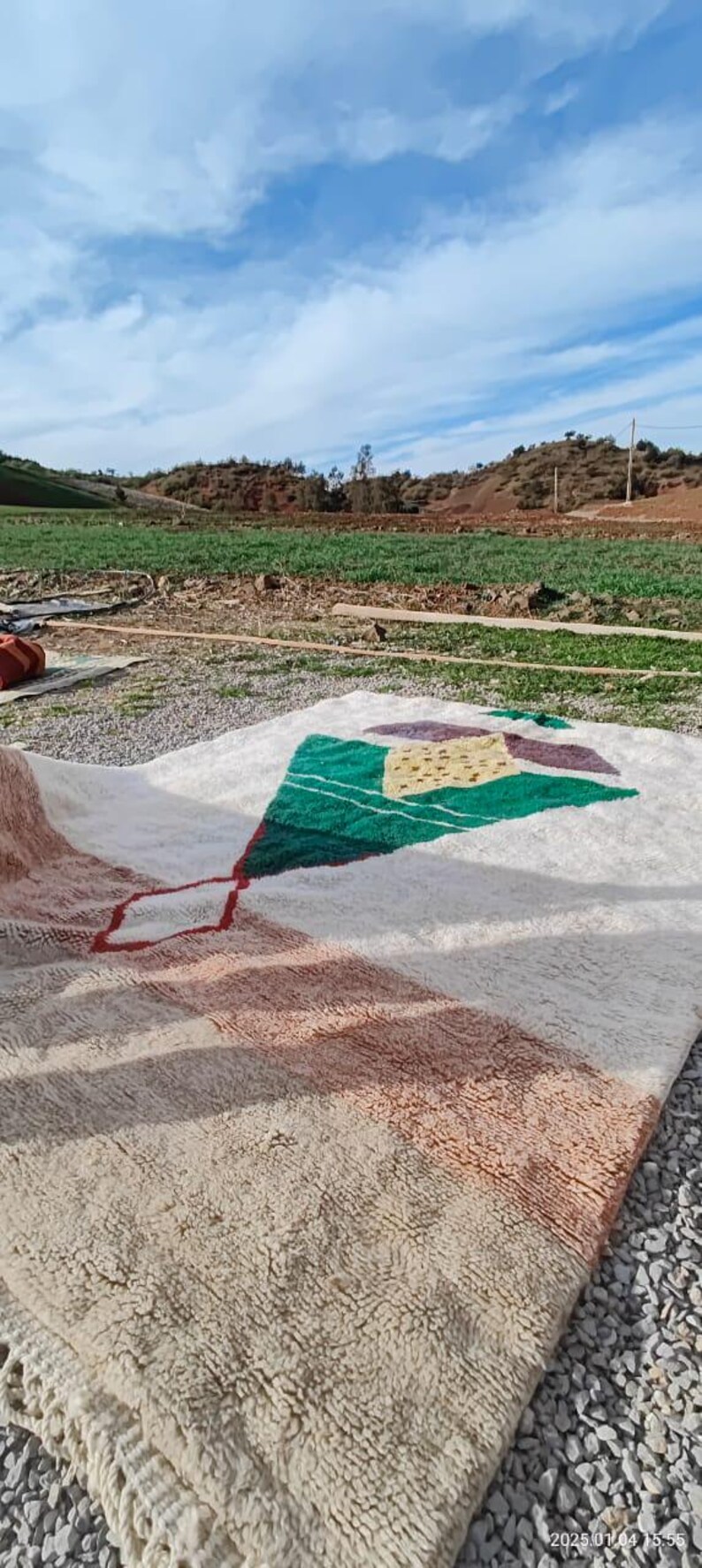 May include: A white, handwoven rug with a geometric design in shades of green, brown, and red. The rug is laid out on a gravel surface with a green field and blue sky in the background.