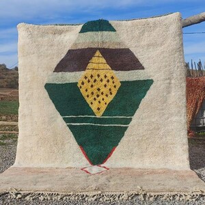 May include: A white wool rug with a geometric design in shades of green, brown, and yellow. The design features a large diamond shape with a smaller diamond inside.