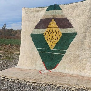 May include: A white rug with a geometric design in shades of green, brown, and yellow. The design features a large diamond shape with smaller diamonds inside.