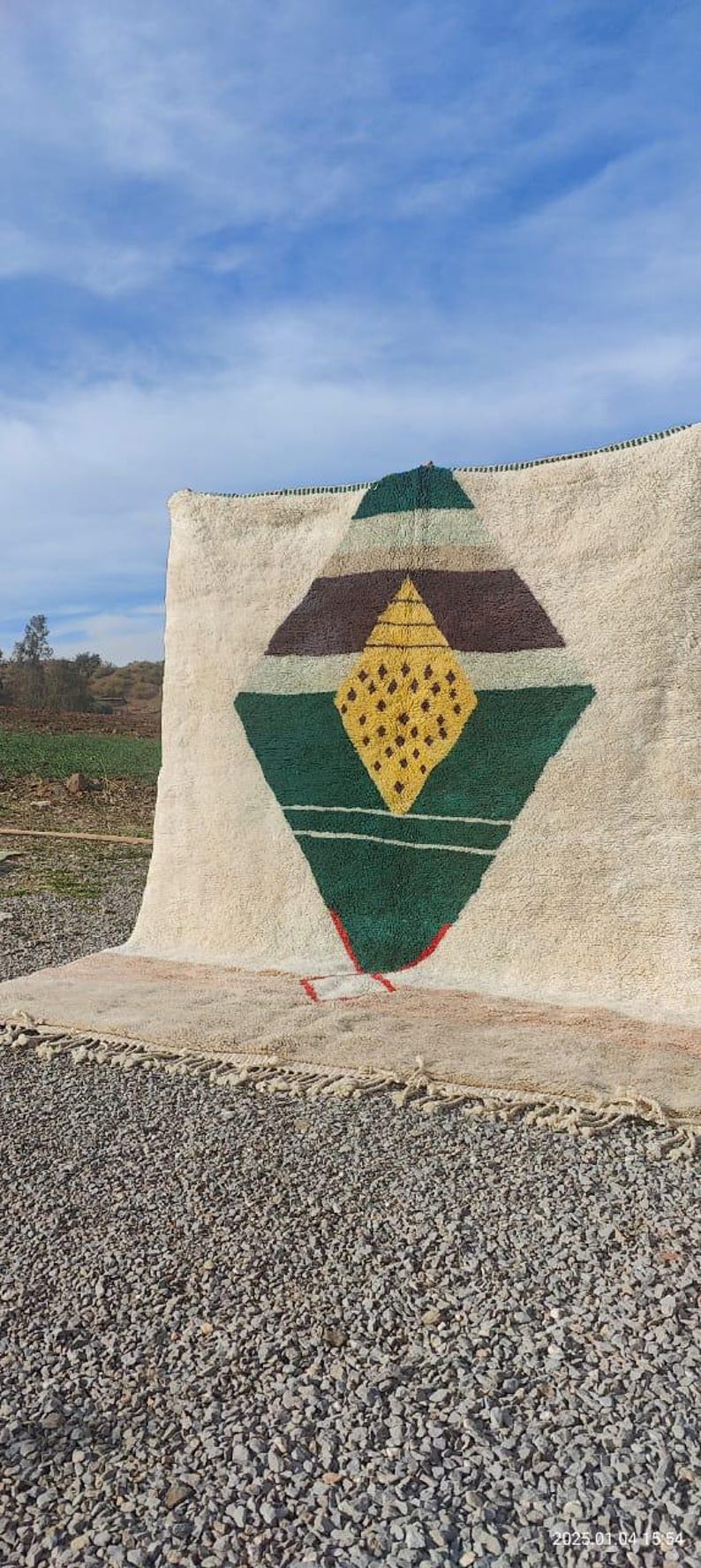 May include: A white rug with a geometric design in shades of green, brown, and yellow. The design features a large diamond shape with smaller diamonds inside.