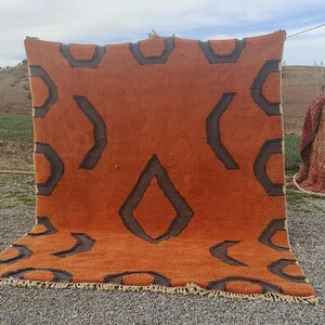 May include: A large, orange and brown patterned rug with a geometric design. The rug is hanging on a clothesline outside, with a view of a field and sky in the background.