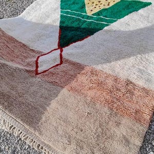 May include: A white, beige, green, and red Moroccan rug with a geometric design. The rug has a fringe border.