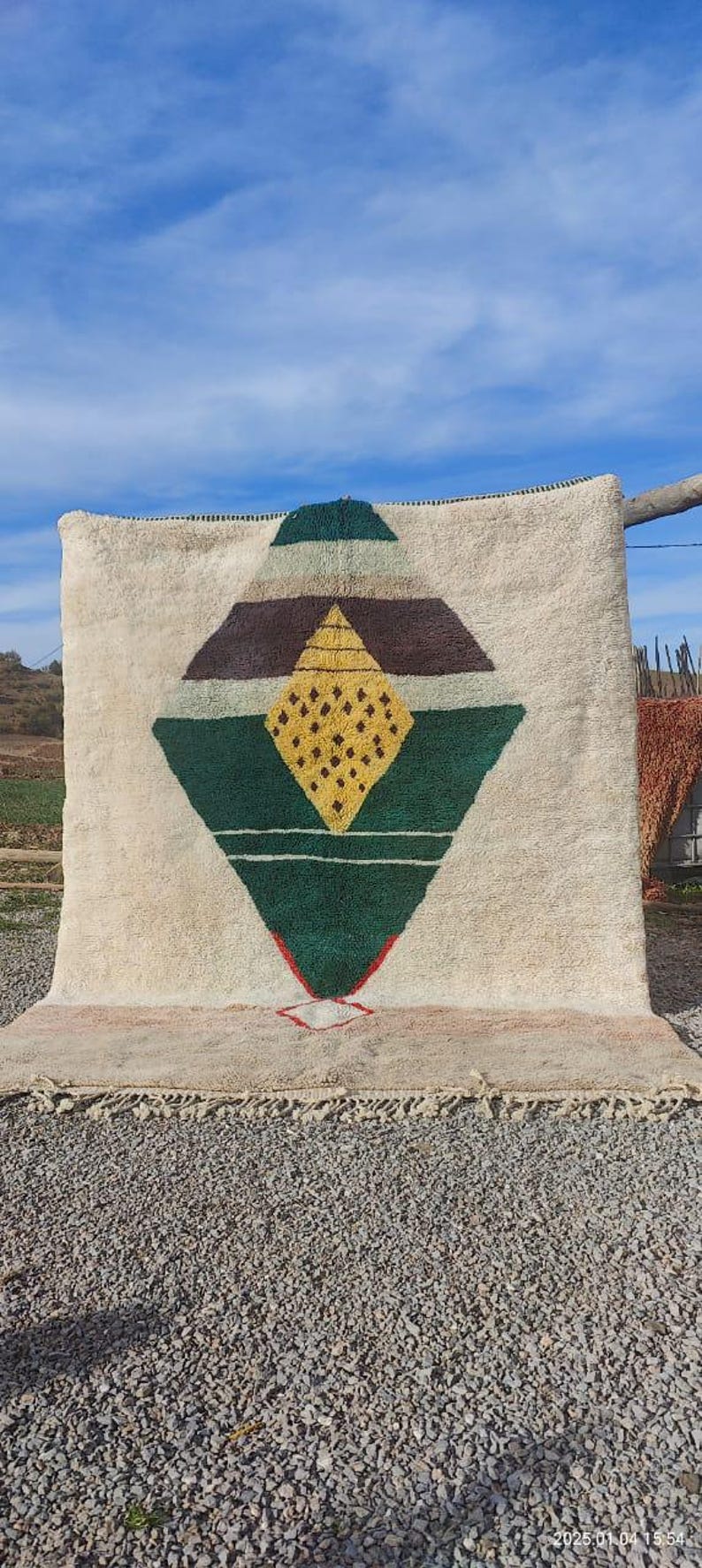 May include: A white wool rug with a geometric design in shades of green, brown, and yellow. The design features a large diamond shape with a smaller diamond inside.