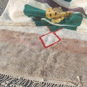 May include: A white, beige, brown, green, and yellow rug with a red square design. The rug has a fringe border and is lying on a bed of gray gravel.