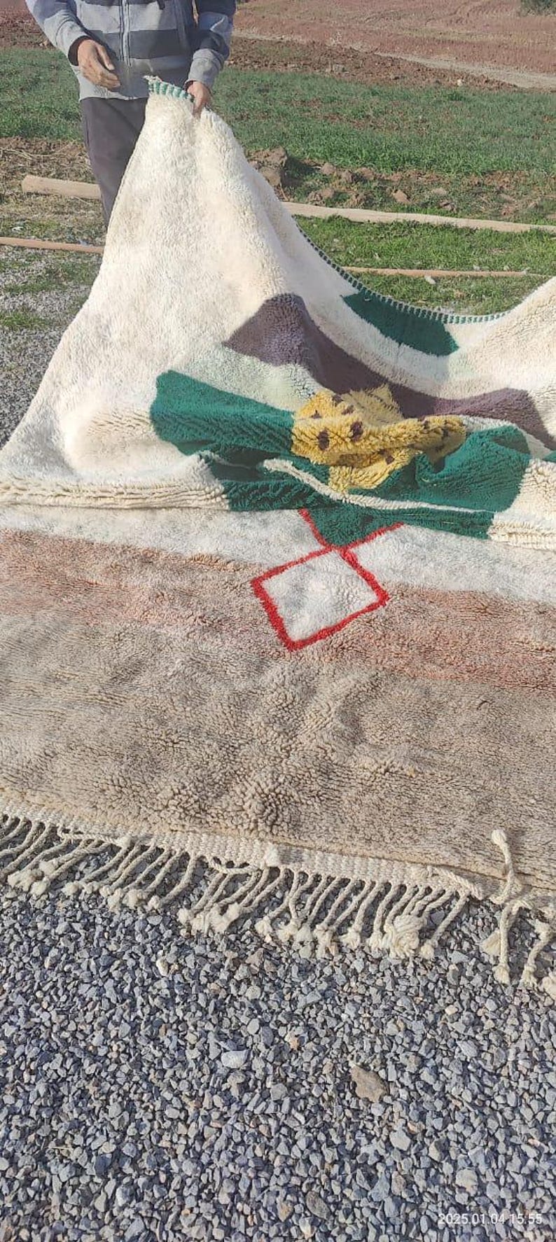 May include: A white, beige, brown, green, and yellow rug with a red square design. The rug has a fringe border and is lying on a bed of gray gravel.
