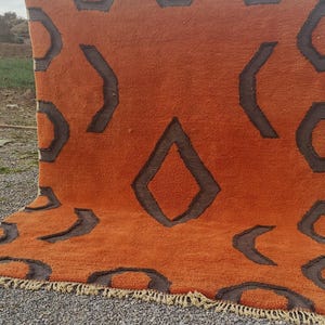 May include: A large, orange rug with a geometric pattern in brown. The rug has a fringe border.
