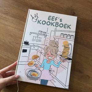 Op de afbeelding: Een kookboek getiteld "EEF's KOOKBOEK" met een cartoonillustratie van een vrouw die pannenkoeken bakt. De omslag is wit met roze en blauwe accenten. Het boek wordt door een hand op een houten oppervlak gehouden.