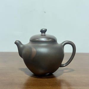 May include: A dark brown, matte-finish clay teapot with a rounded body, a curved spout, a matching lid with a small knob handle, and a C-shaped handle. The surface of the teapot features subtle, irregular color variations in a muted, earthy tone.