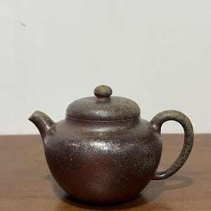 May include: A small, round, dark brown teapot with a textured, speckled surface. It features a curved handle, a short spout, and a matching lid with a small knob on top. The finish has a rustic, weathered appearance with subtle metallic highlights.