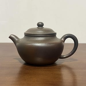 以下が含まれることがあります： マットな質感のダークブラウンの陶器製ティーポット。丸みを帯びた本体、曲線を描く注ぎ口、円形の持ち手、そして上部に小さな丸い取っ手が付いた蓋で構成されています。ティーポットは滑らかで均一な質感をしており、シンプルで機能的なデザインです。