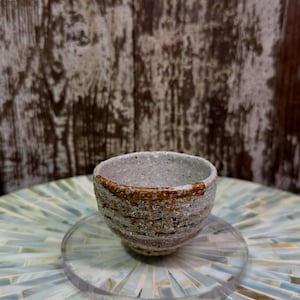 Peut inclure: Une petite tasse en céramique ronde avec une surface rugueuse et texturée dans des tons gris, beige et marron. Le bord présente une bande distincte et irrégulière de glaçure brun rouille, tandis que le corps affiche des stries horizontales.