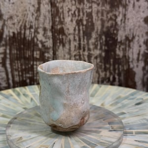 May include: A small, hand-formed ceramic cup with an irregular, organic shape. The exterior features a pale, crackled glaze in shades of off-white and light blue, revealing patches of raw, textured, earthy brown clay near the base and rim.