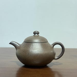 May include: A small, round, dark brown teapot with a textured, matte finish. The teapot features a curved spout, a circular handle, and a matching lid with a small, rounded knob on top. The body has a wide, bulbous shape and a smooth, uniform color.