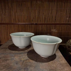 May include: Two pale celadon green ceramic bowls with a subtle scalloped rim design. Each bowl features a smooth, rounded body and a small circular foot base. The glaze has a soft, light-reflecting finish with faint crackle patterns visible.
