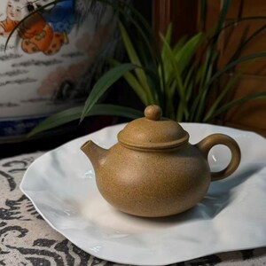 May include: A small, round, tan-colored clay teapot with a textured, speckled surface. The teapot features a short, curved spout, a circular handle, and a matching lid with a small, rounded knob on top, resting on a white, wavy-edged plate.