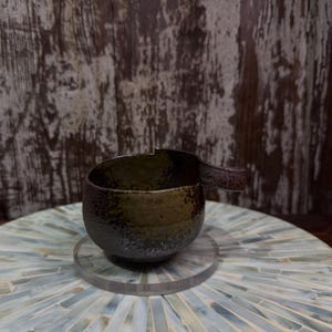 May include: A dark, textured ceramic cup with a mottled brown and black glaze, featuring a single protruding handle on the side and a small notch on the upper rim. The cup has a rustic, uneven surface finish and a deep, rounded bowl shape.