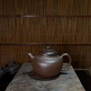 May include: A round, dark brown teapot with a textured, speckled surface. The teapot features a curved handle, a short spout, and a matching lid with a small knob on top. The finish appears matte and earthy, with a consistent dark brown color throughout.