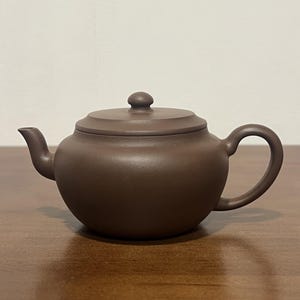 May include: A dark brown, matte-finish clay teapot with a rounded body, a short curved spout, a C-shaped handle, and a flat lid featuring a small, rounded knob handle centered on top. The teapot has a smooth, uniform texture throughout its surface.
