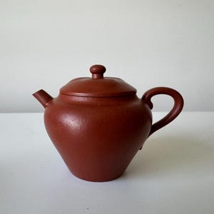 May include: A small, reddish-brown ceramic teapot with a matching lid. The teapot has a rounded body, a curved handle, and a spout. The lid has a small knob on top. The teapot is sitting on a white surface.
