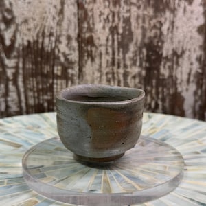 以下が含まれることがあります： 素朴で不揃いな縁を持つ、手作りの小さな陶器のカップ。外側は片面が質感のあるマットなグレーで、もう片面は温かみのあるアースカラーのブラウンになっています。カップには短く頑丈な台座があり、口が広く開いています。