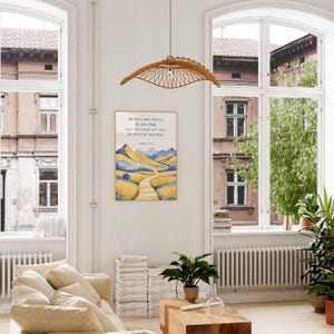 May include: A framed watercolor print with a landscape of yellow fields and blue mountains, with text in German. The print hangs on a white wall in a bright room with large windows. A woven rattan pendant light hangs above a wooden coffee table.