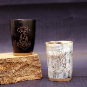 May include: Two Viking-style drinking horns. One is black with a carved Thor's hammer design, the other is a natural horn colour with a carved animal design. Both have a wooden base and are displayed on a wooden block.
