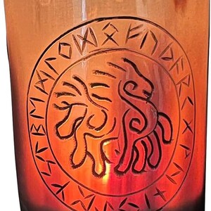 May include: A cylindrical glass candle holder with an etched design. The design features a lion-like figure surrounded by runes. The glass has a gradient of colours, transitioning from a dark brown base to a warm orange at the top.