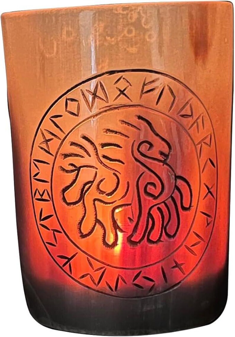 May include: A cylindrical glass candle holder with an etched design. The design features a lion-like figure surrounded by runes. The glass has a gradient of colours, transitioning from a dark brown base to a warm orange at the top.