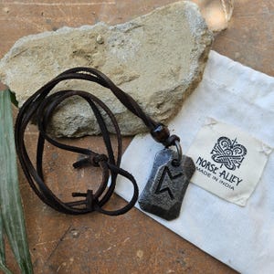 May include: A dark gray pendant necklace with a leather cord. The pendant has a carved rune symbol. A small white bag with the text "NORSE ALLEY MADE IN INDIA" is next to the necklace.