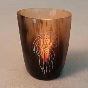 May include: A brown glass votive candle holder featuring a horse design etched on the front. The candle holder has a gradient colour, transitioning from a dark brown at the base to a lighter brown at the top. The horse design is illuminated by the candle inside.
