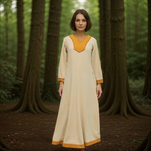 A woman stands in a forest, wearing a long, cream-colored dress with a yellow V-neck collar and yellow trim on the cuffs and hem.
